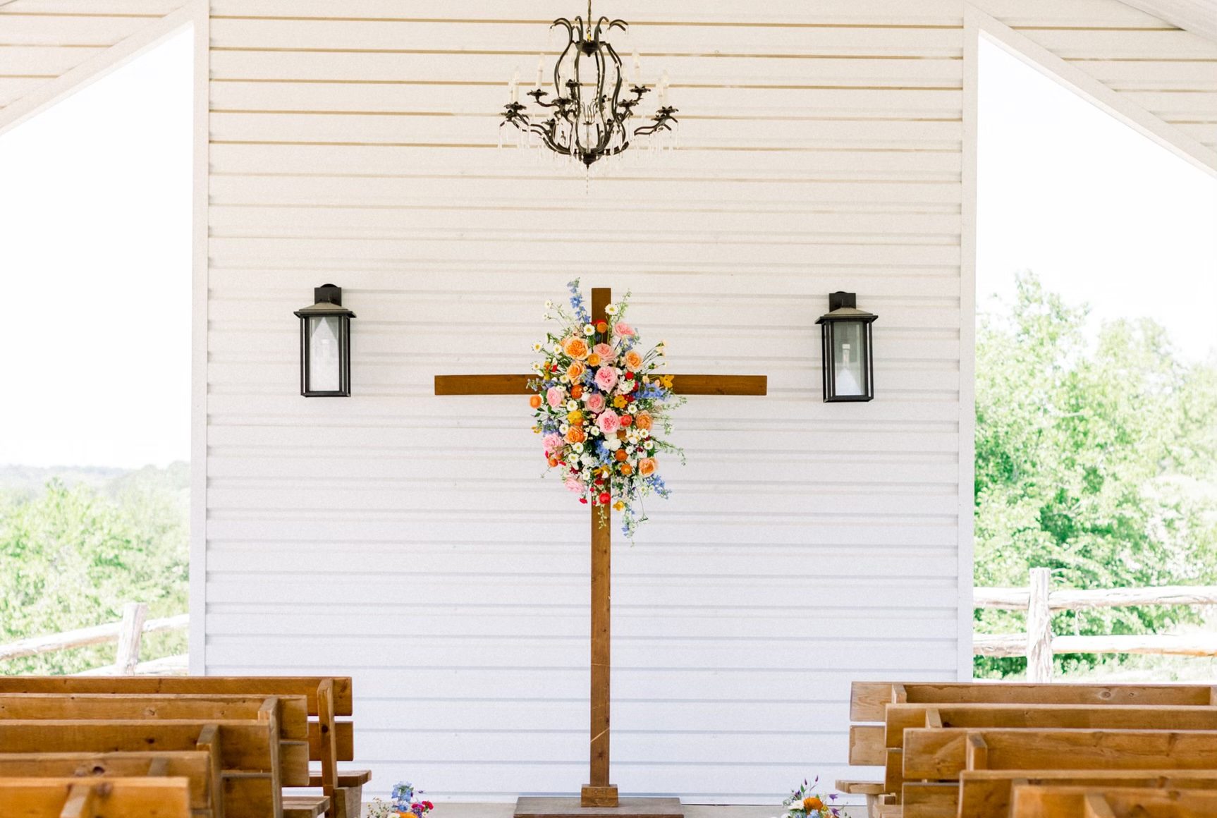 Inside the white open-air chapel at Diamond H3 Ranch, a wooden cross with a colorful floral arrangement above wooden pews, open to the hill-country views